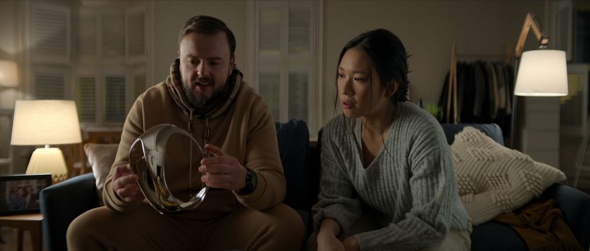 John Bradley dans le rôle de Jack Rooney et Jess Hong dans le rôle de Jin Cheng sont assis sur un canapé et regardent le casque VR chromé dans l'épisode 102 de 3 Body Problem