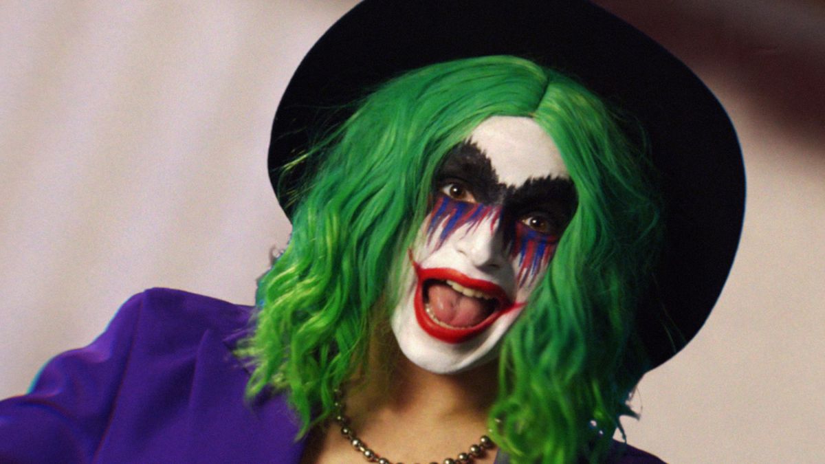 Vera Drew dans le rôle de Joker l'Arlequin, un mashup de Joker et Harley Quinn, en perruque verte, maquillage de clown avec un grand sourire rouge à lèvres, une veste de costume violette et un chapeau noir dans The People's Joker