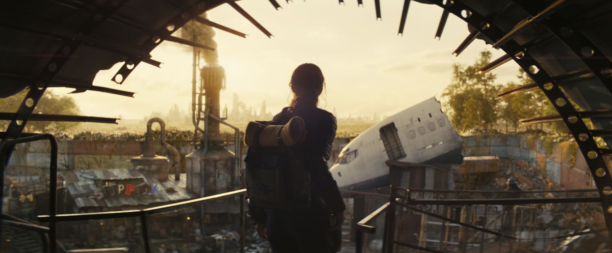 Une jeune femme avec un sac à dos regardant une ville faite de métal récupéré dans la série télévisée Fallout.