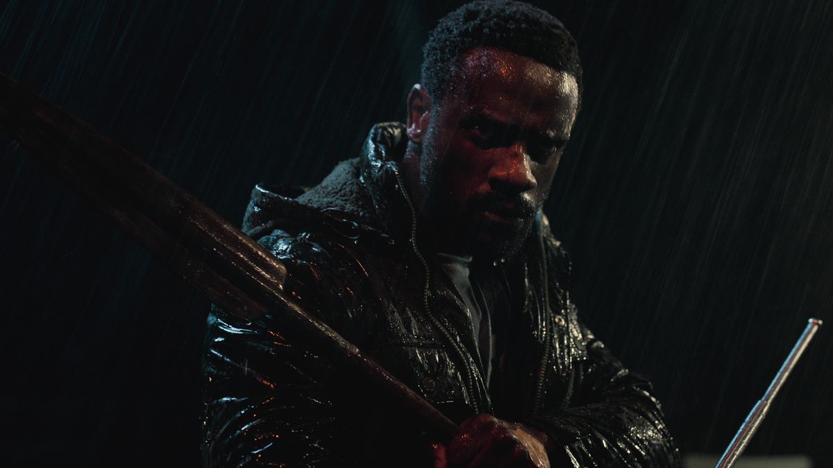 Un homme vêtu d’une veste en cuir debout sous la pluie tenant des armes.