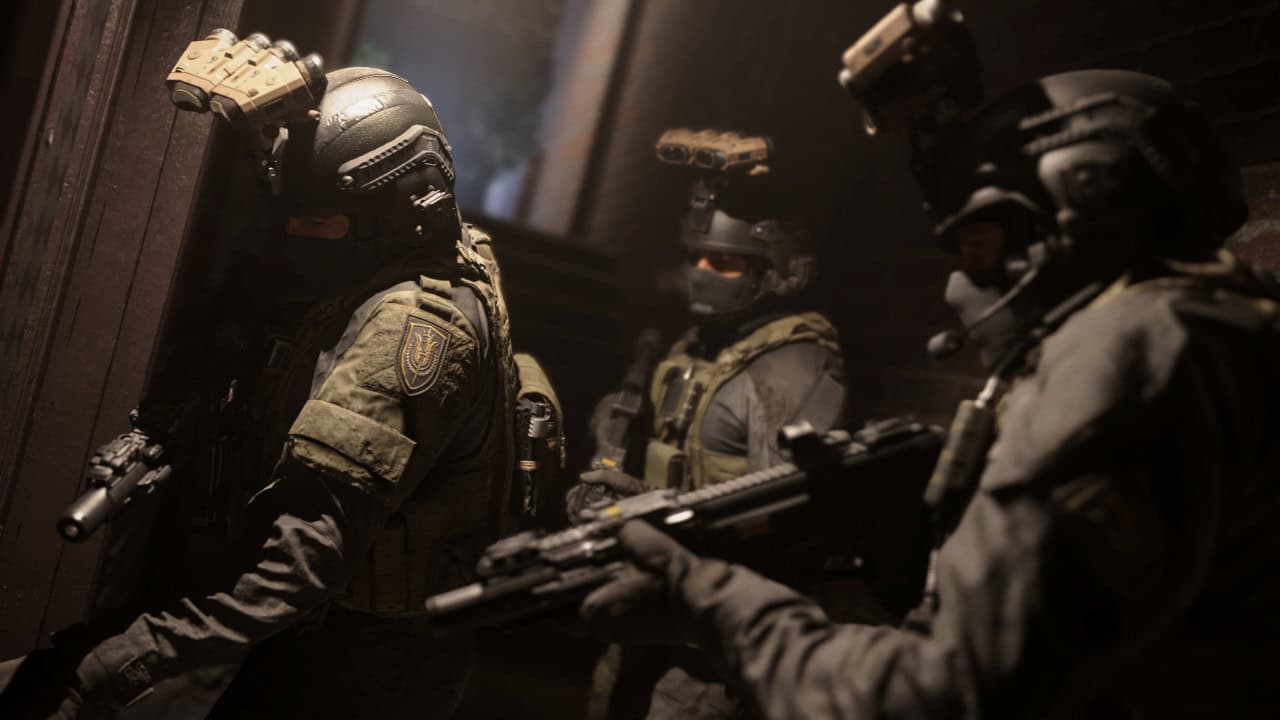 Une équipe des forces spéciales en tenue tactique se prépare à percer un bâtiment, tenant des armes, éclairée par une faible lumière filtrant à travers la fumée dans le jeu visuellement époustouflant MW 2019.