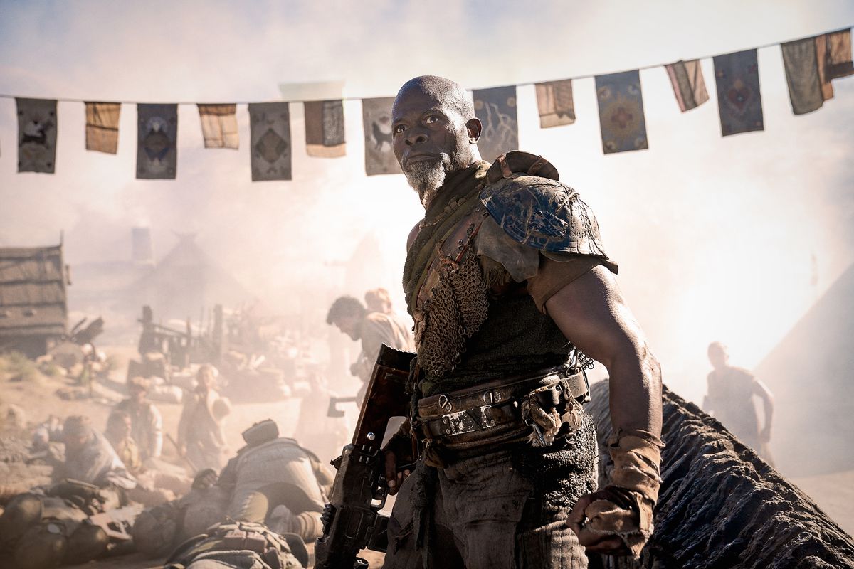 Djimon Hounsou dans le rôle du général Titus dans Rebel Moon – Deuxième partie : Le Scargiver tenant une arme à ses côtés alors qu'il se tient près d'un village massacré