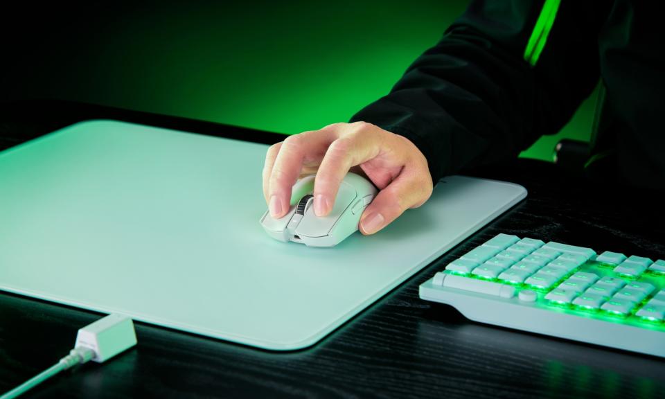 Une souris de jeu Razer Viper V3 Pro blanche est placée sur un tapis de souris blanc sur un bureau noir, avec un clavier blanc brillant d'un éclairage vert situé sur le côté.