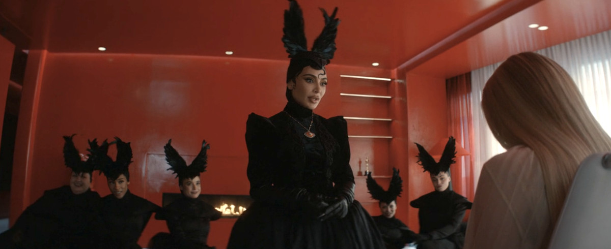 Kim Kardashian dans une coiffure à plumes noires criardes se moque d'Emma Roberts.  Derrière elle se trouve un troupeau de femmes portant la même coiffe.  Ils se trouvent tous dans un loft moderne rouge vif. 
