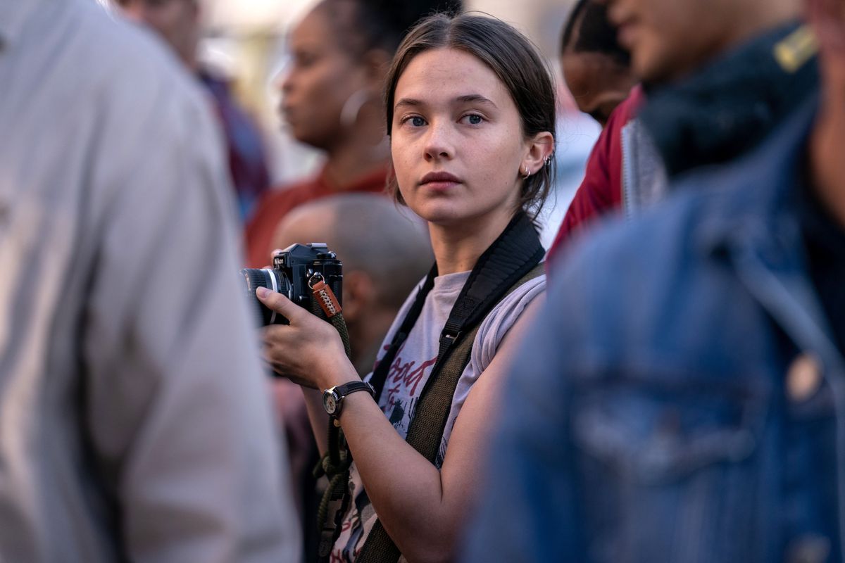 Jessie (Cailee Spaeny), une jeune photojournaliste tenant un appareil photo, se tient au milieu d'une foule dans Civil War d'Alex Garland.