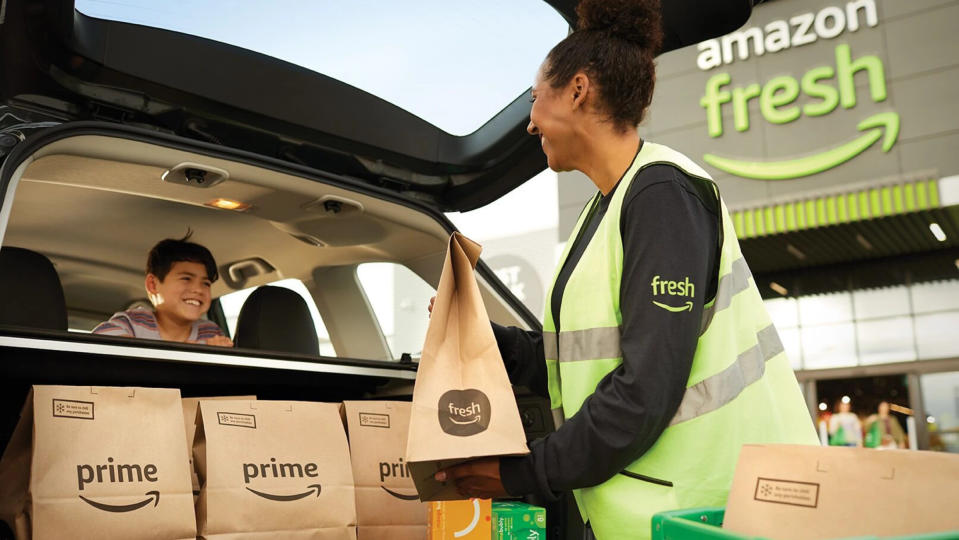 Un employé d'Amazon Fresh vêtu d'un gilet vert fluo charge des produits d'épicerie Prime Fresh dans une voiture à hayon avec un enfant souriant sur la banquette arrière.  La personne qui charge les courses sourit en retour. 