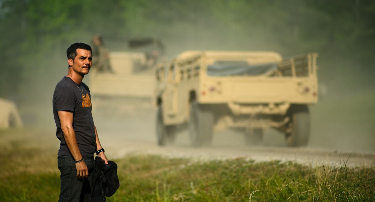 Le correspondant de guerre Joel (Wagner Moura) se tient dans l'herbe au bord d'une route poussiéreuse alors que d'énormes véhicules militaires passent dans la guerre civile d'Alex Garland.