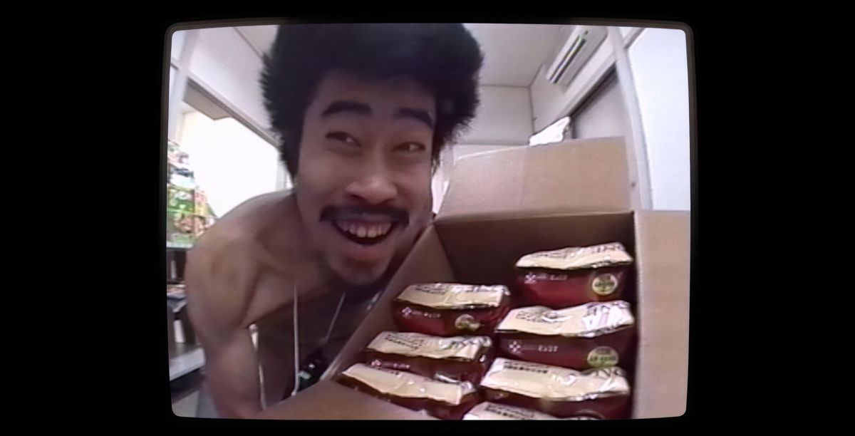 Le comédien japonais Nasubi, nu et avec un énorme pouf aux cheveux sauvages, sourit en brandissant une boîte pleine de sacs de nourriture pour chien qu'il s'apprête à manger dans le jeu télévisé Susunu !  Denpa Shōnen, comme on le voit dans le documentaire The Contestant