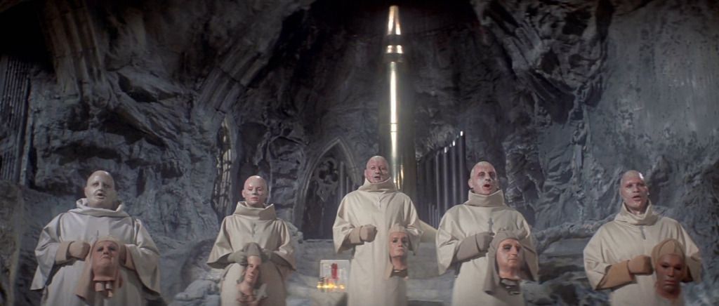 Un groupe de personnages chauves vêtus de costumes de moine et tenant des têtes de mannequin dans une caverne avec un grand intérieur semblable à une église dans Sous la planète des singes.