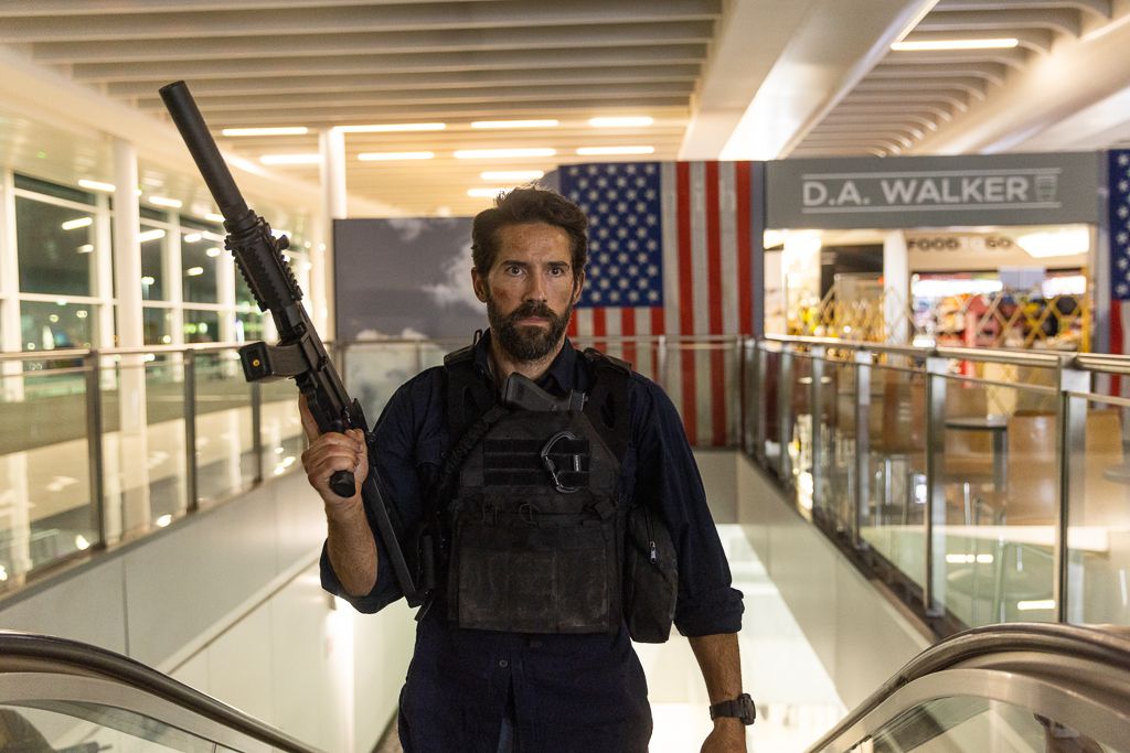 Scott Adkins porte un gilet pare-balles et tient une arme à feu en montant un escalier roulant à l'aéroport dans One More Shot.