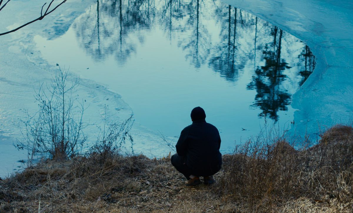Un personnage en manteau et bonnet vu accroupi de dos au bord d'un lac à moitié gelé, les bois se reflétant à sa surface dans le film Evil Does Not Exist.