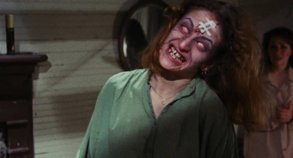 Une jeune femme commence à se transformer en Deadite, les yeux blancs, avec un sourire malicieux sur le visage, dans The Evil Dead.