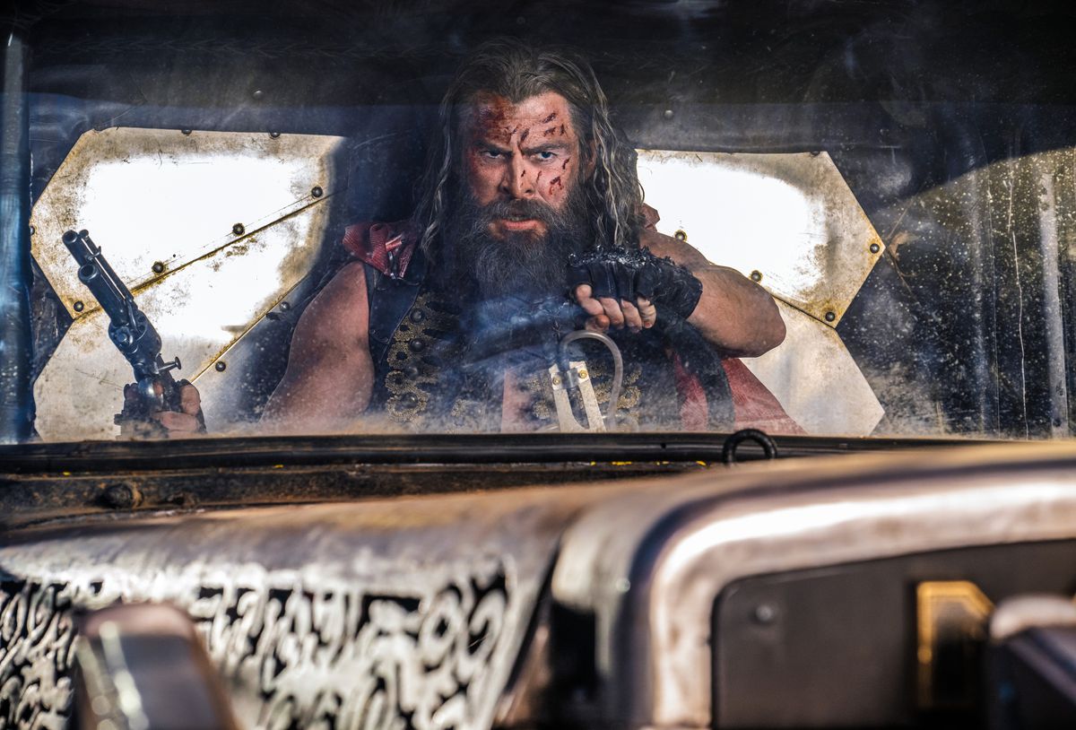 Dementus (Chris Hemsworth), un guerrier des Terres désolées aux cheveux et à la barbe hirsute, tient un pistolet dans une main et dirige un véhicule de l'autre dans Furiosa de George Miller.