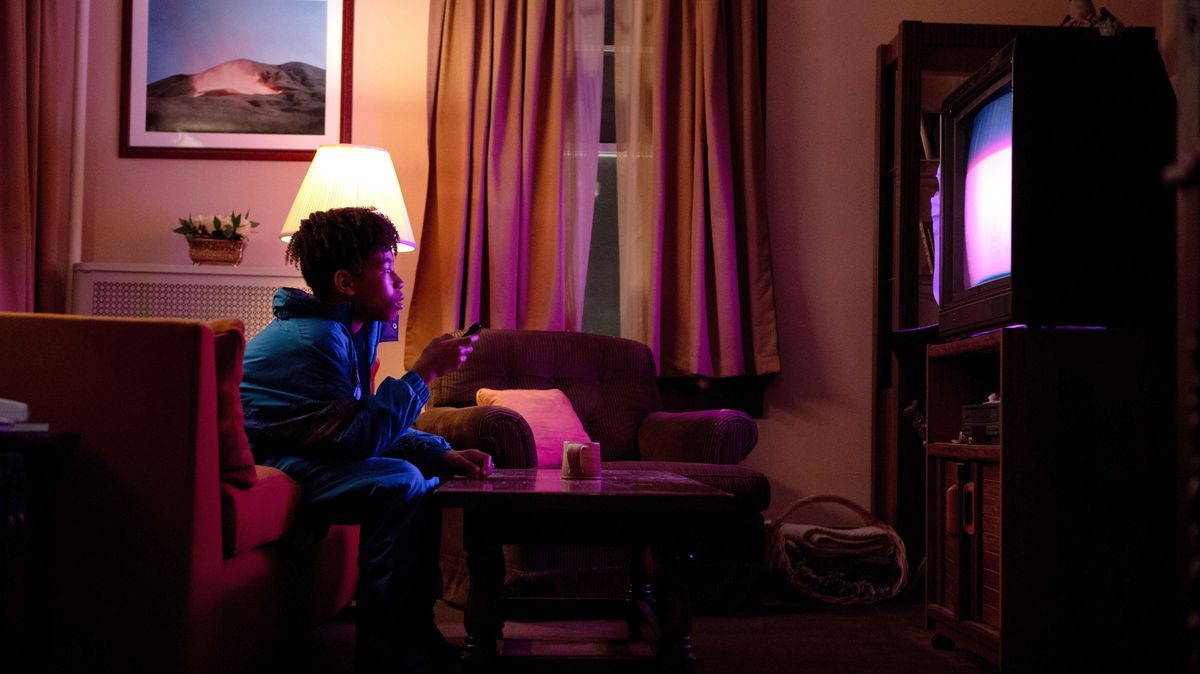 Le jeune Owen (Ian Foreman) est assis sur un canapé rouge dans un salon sombre, regardant la télévision, dans I Saw the TV Glow de Jane Schoenbrun.