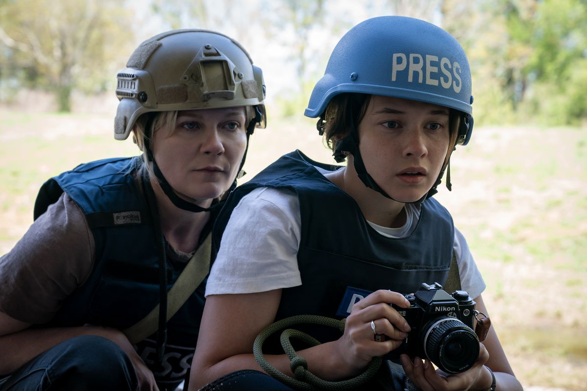 Les photojournalistes Lee Miller (Kirsten Dunst) et Jessie (Cailee Spaeny) se serrent les uns contre les autres, Jessie tenant son appareil photo et portant un casque bleu vif sur lequel est écrit « PRESS », dans Civil War d'Alex Garland.