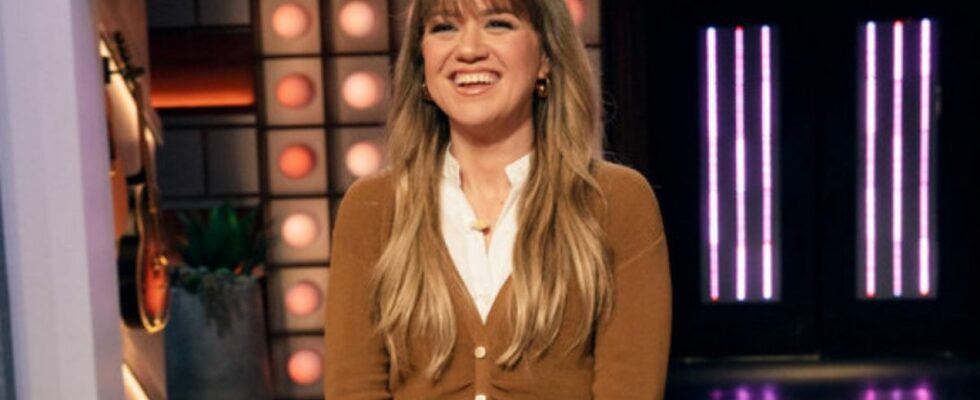 Kelly Clarkson, wearing a white shirt and brown cardigan, smiles at the crowd from the stage of The Kelly Clarkson Show.