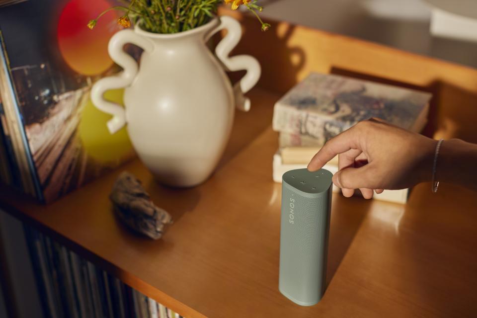 Photo lifestyle pour l'enceinte portable Sonos Roam 2.  Il est assis sur une table de chevet très fréquentée tandis qu'une main atteint le cadre (en partant de la droite) pour toucher ses commandes.
