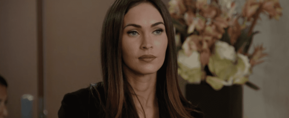 Megan Fox sits in a black blouse in front of a vase of flowers in a scene from New Girl