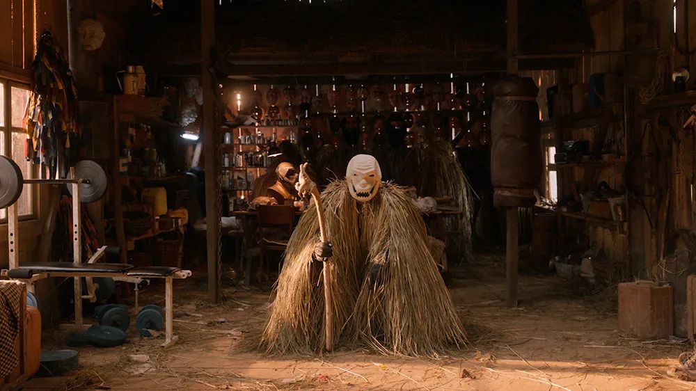 Une personne portant un masque lorgnant et un costume de paille tenant un bâton au milieu d'un atelier à Pamfir.
