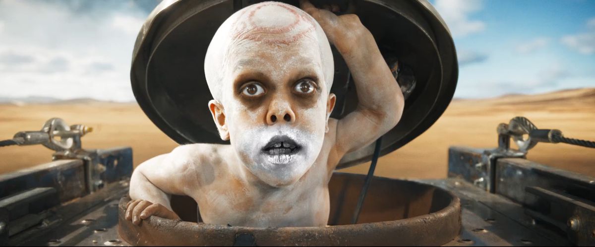 War Pup (Quaden Bayles, torse nu, peint en blanc, avec de la peinture chromée en aérosol sur la bouche et des scarifications circulaires sur le crâne) sort d'une trappe dans le War Rig à Furiosa