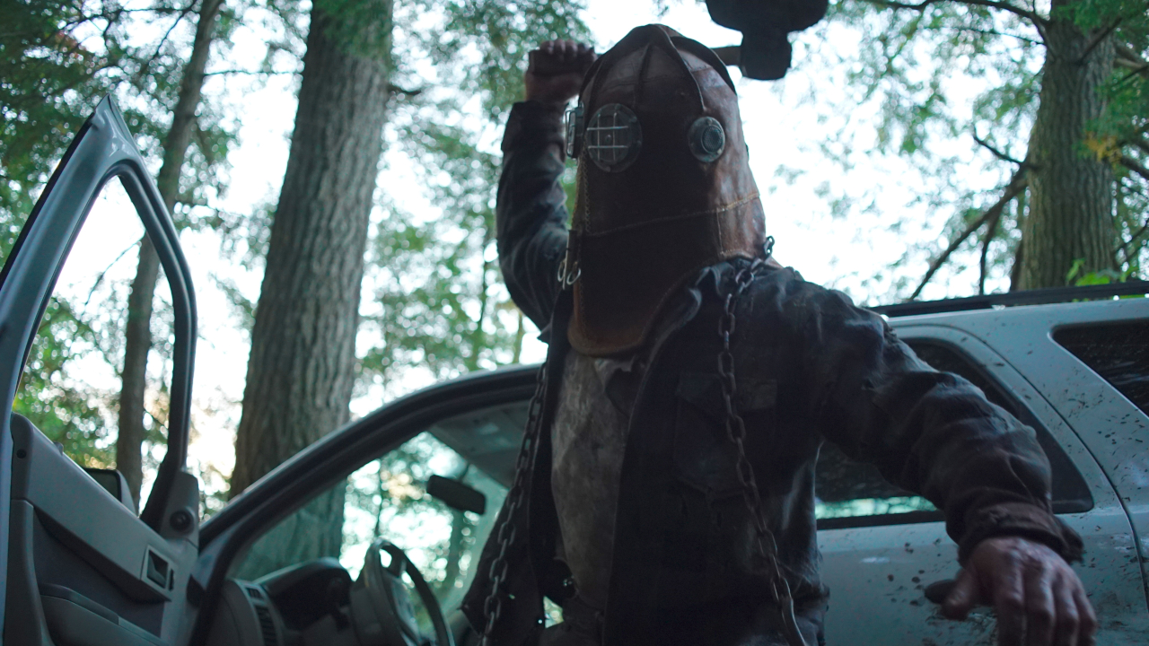 Ry Barrett porte un masque de pompier alors qu'il lève une hache devant une voiture arrêtée dans In A Violent Nature.