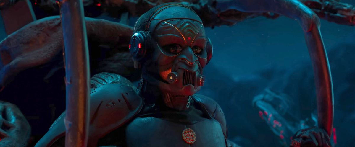 dans une image promotionnelle du blockbuster de science-fiction indien Kalki 2898 AD, une figure humanoïde en armure et masque en métal est assise dans un espace sombre, entourée d'espars métalliques
