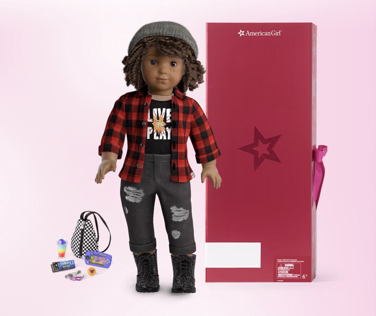 Une poupée American Girl à créer soi-même est présentée portant une flanelle et un jean déchiré.  La poupée a des cheveux bruns courts et bouclés, une peau brune et des taches de rousseur.