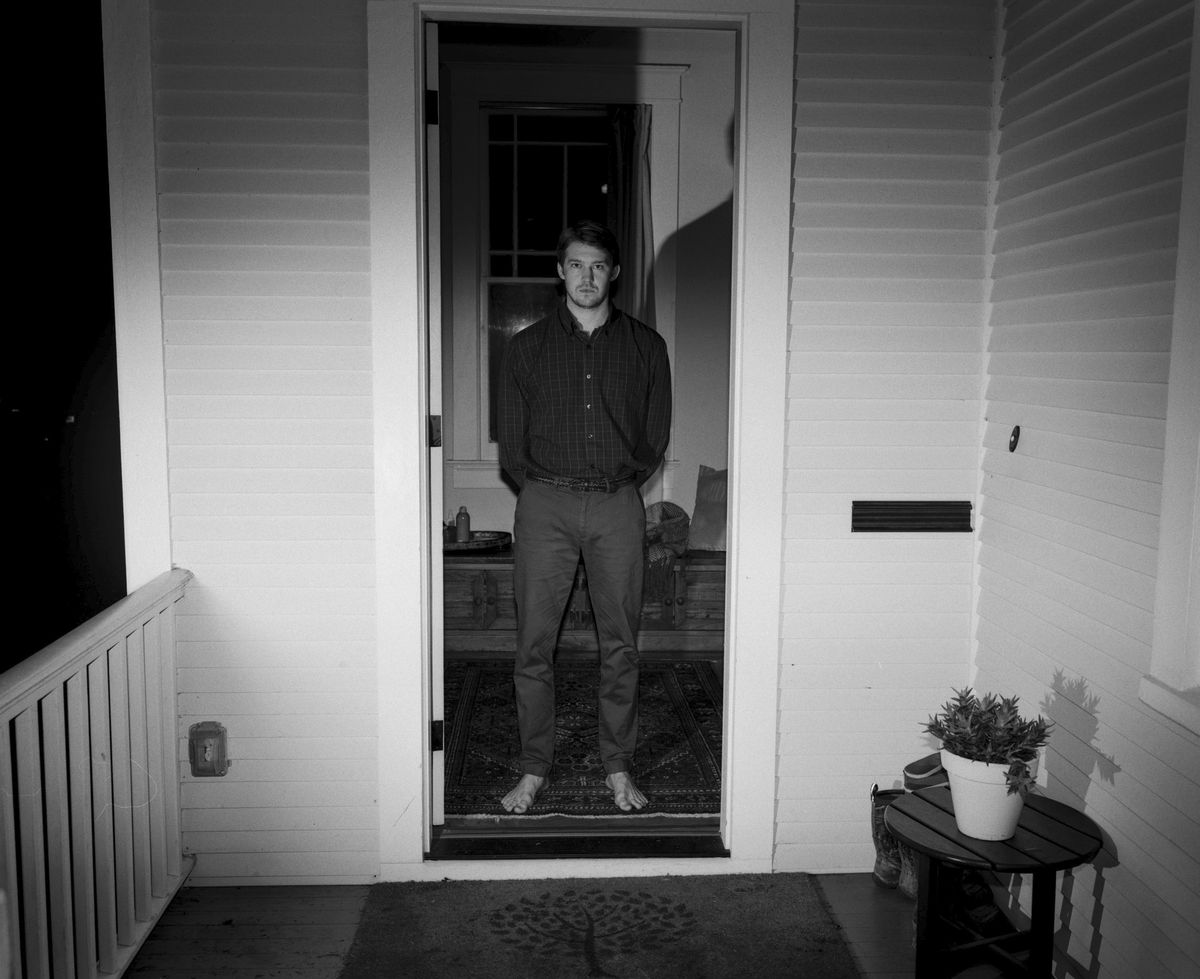 Joe Alwyn, pieds nus et photographié en noir et blanc, se tient sur le pas de la porte d'une maison dans Kinds of Kindness