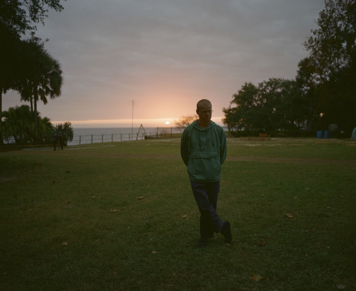 Jesse Plemons, avec une coupe buzz et portant un coupe-vent bleu sarcelle, se tient dans un jardin au bord de la mer au coucher du soleil dans Kinds of Kindness