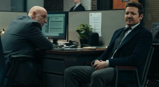 Hugh Dillon sitting in front of a computer looking at Jeremy Renner who is sitting by his desk facing forward.