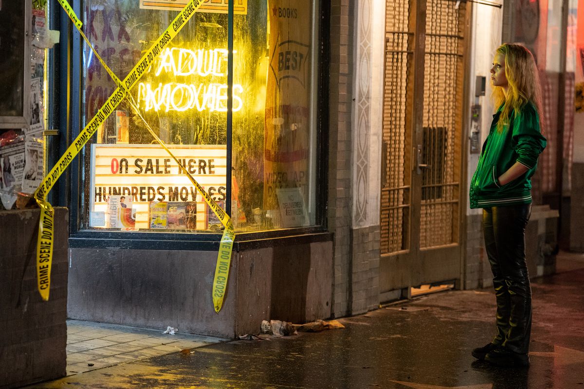 Maxine (Mia Goth) se tient devant un magasin avec un panneau jaune fluo « films pour adultes » et un ruban de police « scène de crime à ne pas traverser » accroché en X sur la porte dans Maxxxine de Ti West