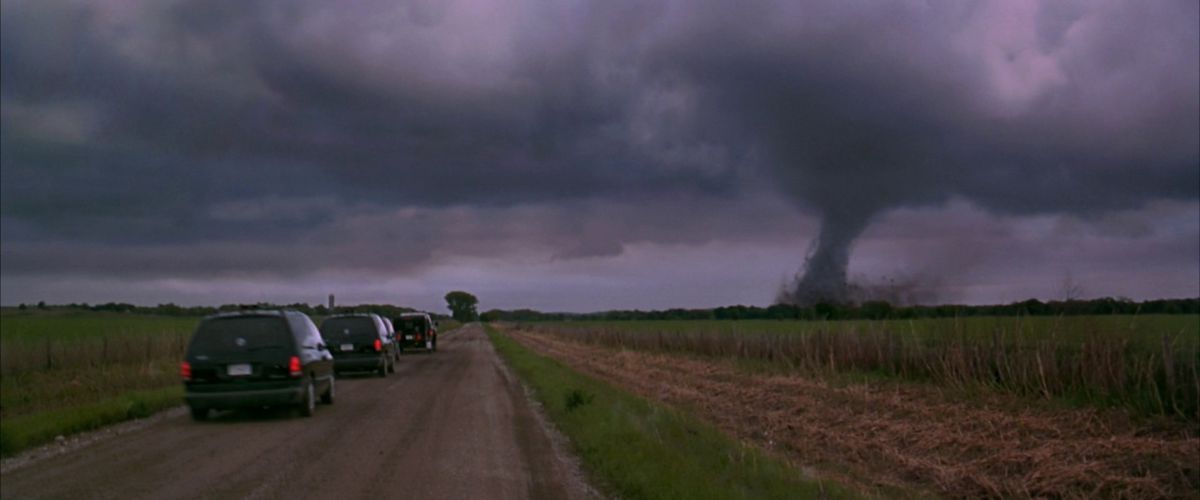 Trois camionnettes roulent sur une route de campagne avec une grande tornade visible au loin dans Twistter.