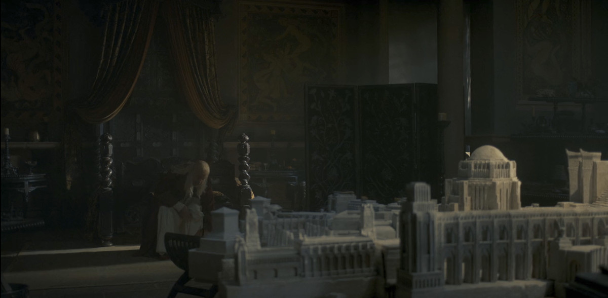 Le roi Viserys (Paddy Considine) assis dans sa chambre, avec sa maquette de Port-Réal devant lui. On peut voir des fresques de sexe en arrière-plan derrière lui sur le mur de sa chambre