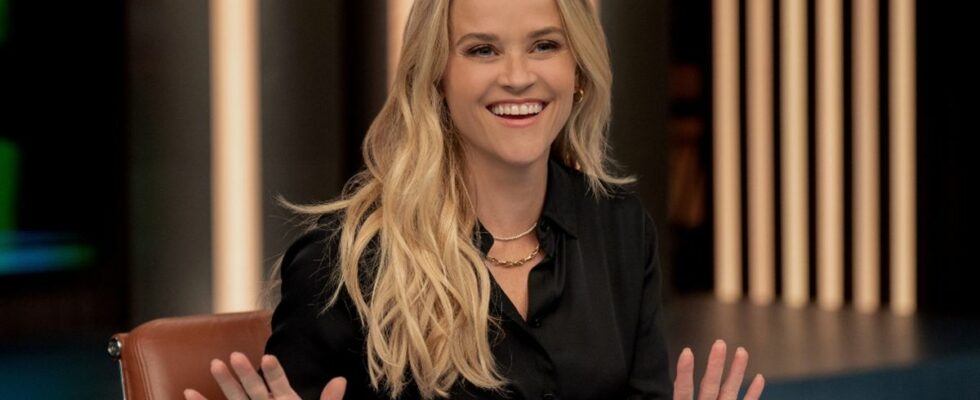 A press image of Reese Witherspoon sitting behind a desk smiling with her hands slightly lifted off the table in Season 3 of The Morning Show.