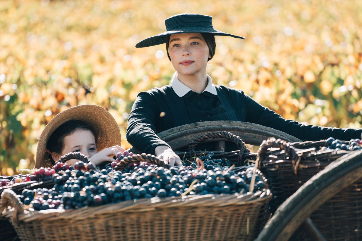 Une femme en robe noire et chapeau debout à côté d'un panier de raisins devant un champ de maïs dans Veuve Clicquot.