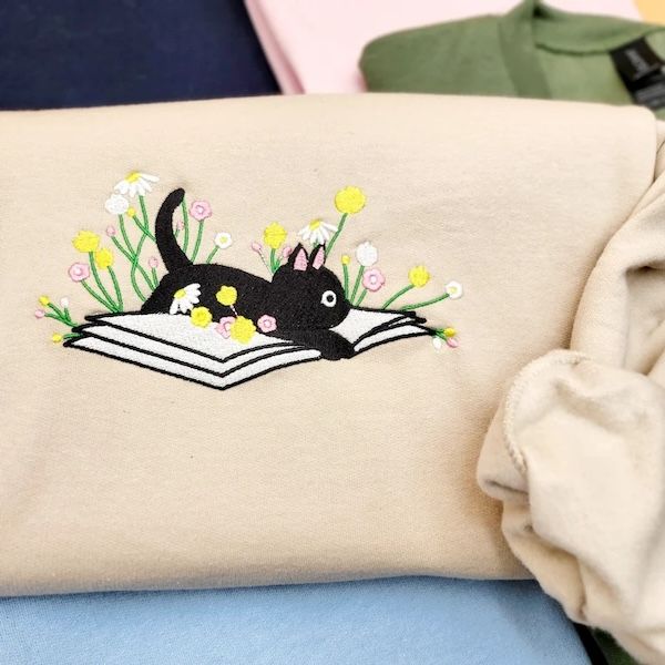 pull ras du cou crème avec un motif brodé d'un chat noir et de fleurs reposant à l'intérieur d'un livre