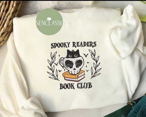 pull ras du cou crème avec texte noir sur le devant qui dit "Club de lecture Spooky Readers." le texte est organisé autour d'un graphique représentant un petit chat noir avec de grands yeux à l'intérieur d'une tasse en forme de crâne sur une pile de livres