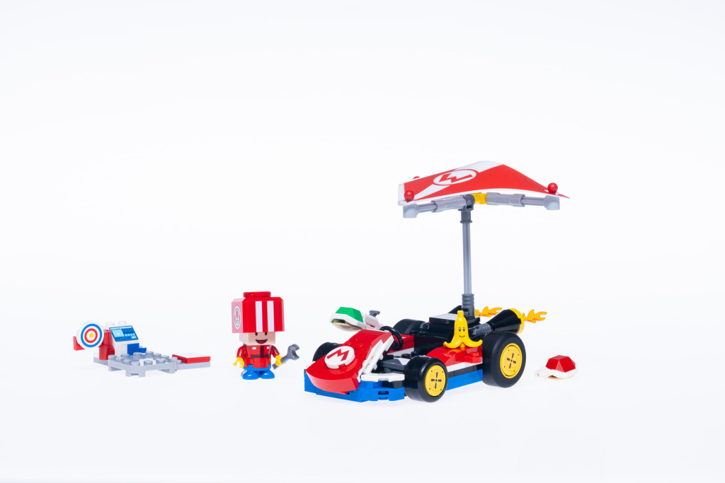 Images promotionnelles officielles de l'ensemble LEGO Mario Kart Standard Cart. On y voit un Mario Go-Kart standard construit en briques LEGO avec un parapente. Sur le capot se trouvent une coque verte et une peau de banane prêtes à tirer sur les adversaires. Il y a une coque rouge près du pneu arrière. À côté du kart se trouve un personnage Toad construit en briques tenant une clé à molette à côté d'un ordinateur