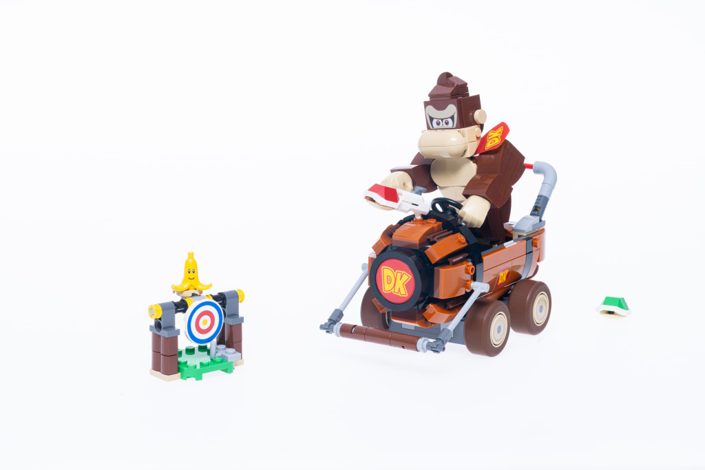 Images promotionnelles officielles pour le set LEGO Mario Kart Donkey Kong et DK Jumbo. On y voit un Donkey Kong construit en briques LEGO assis dans son kart DK. Il tient une carapace rouge dans sa main comme pour la lancer sur une cible à sa droite. Derrière le kart se trouve une carapace verte.