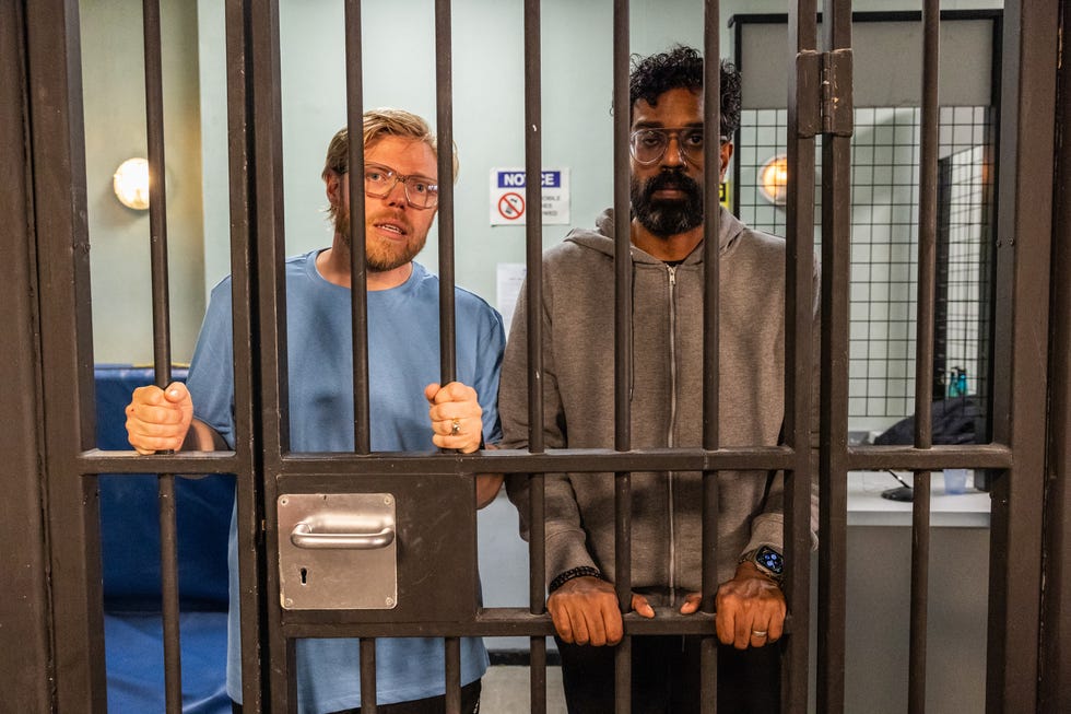 Rob Beckett et Romesh Ranganathan se tiennent derrière une porte de prison à barreaux alors qu'ils tournent pour Hollyoaks