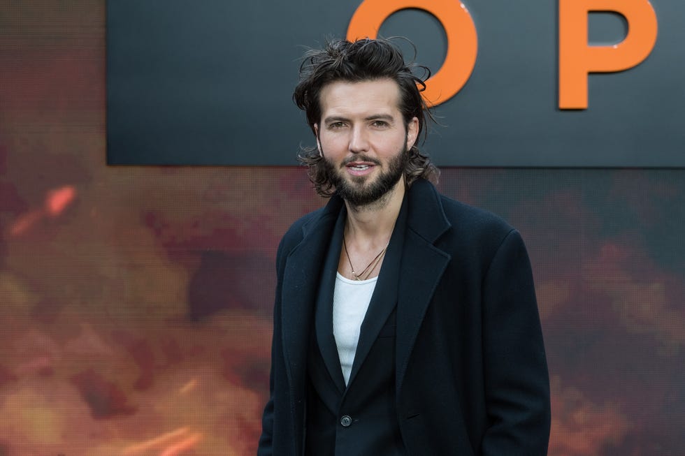 Guy Burnet, un jeune homme regarde la caméra, il a les cheveux noirs jusqu'aux épaules et une barbe, il porte un t-shirt blanc avec un blazer noir