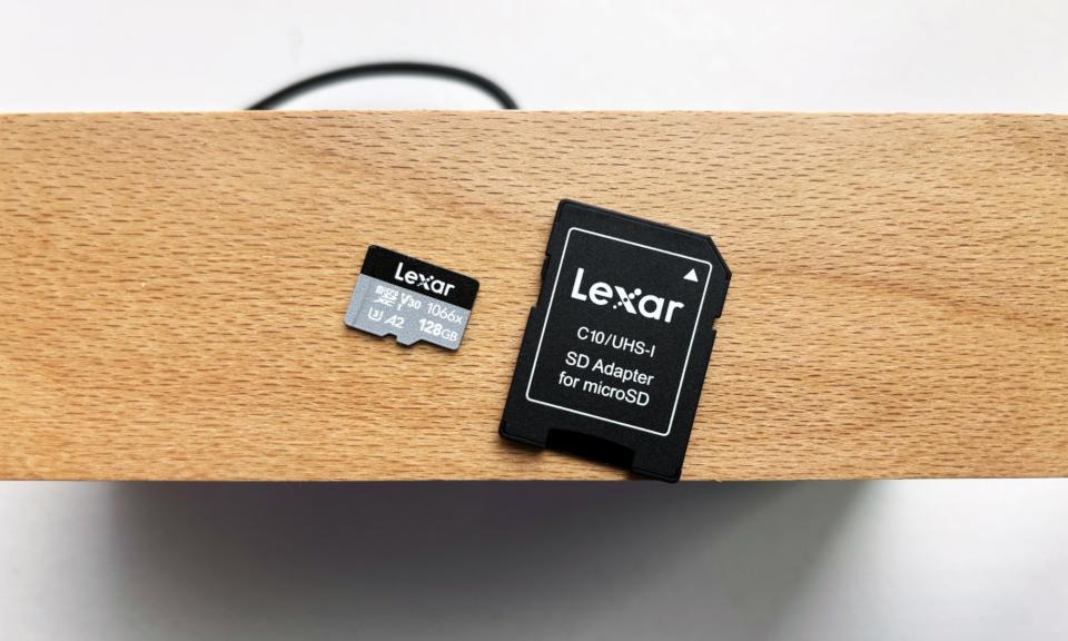 Une carte microSD Lexar Professional 1066x argentée et noire et son adaptateur de carte SD noir reposent sur une étagère en bois marron au-dessus d'un rebord de fenêtre blanc.