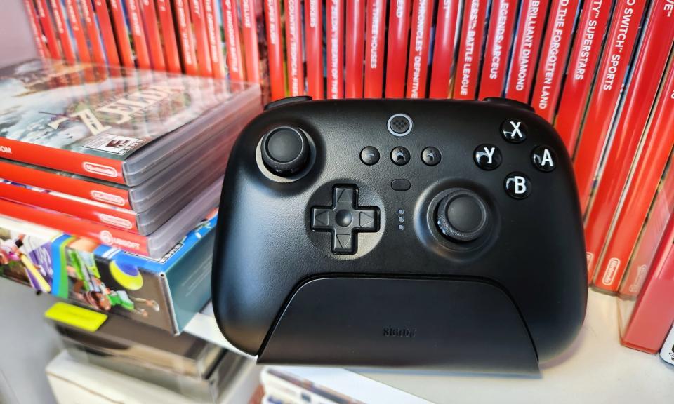 Un contrôleur de jeu vidéo noir, le contrôleur Bluetooth 8BitDo Ultimate, repose sur une étagère blanche devant une rangée de plusieurs boîtiers de jeux vidéo Nintendo Switch rouges.