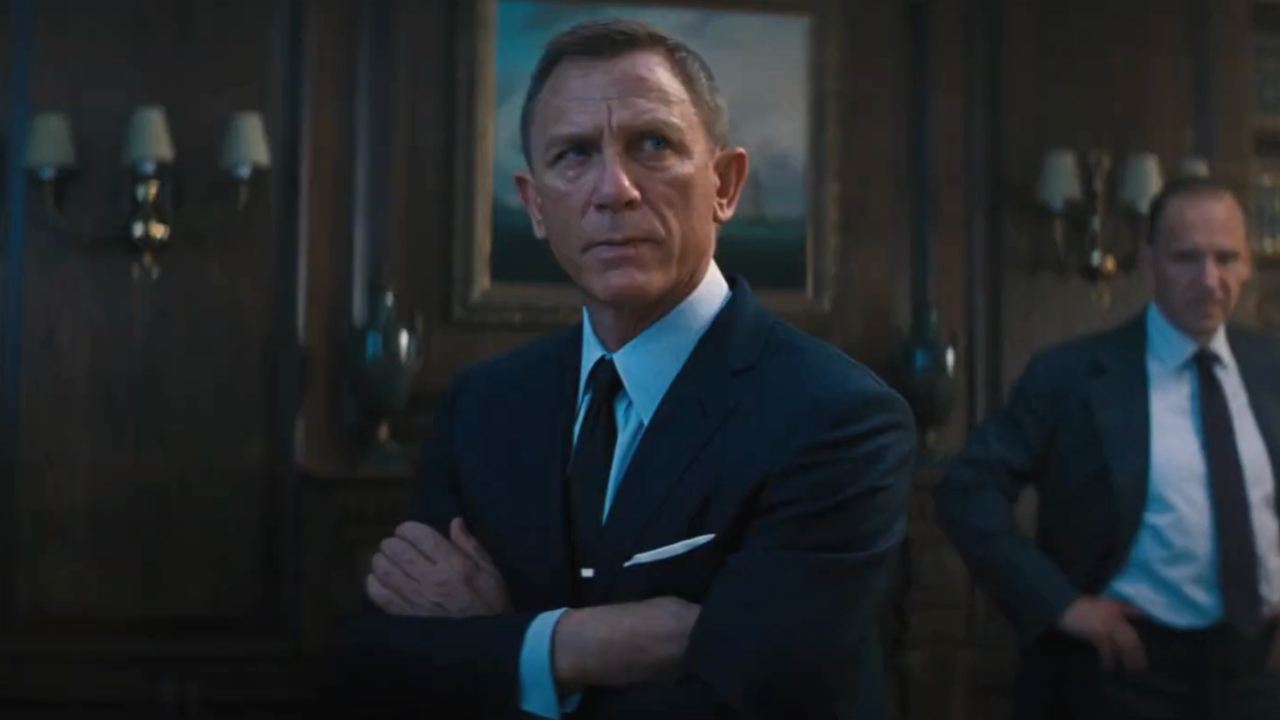 Daniel Craig est assis, les bras croisés, l'air plutôt bouleversé pendant que Ralph Fiennes regarde No Time To Die.